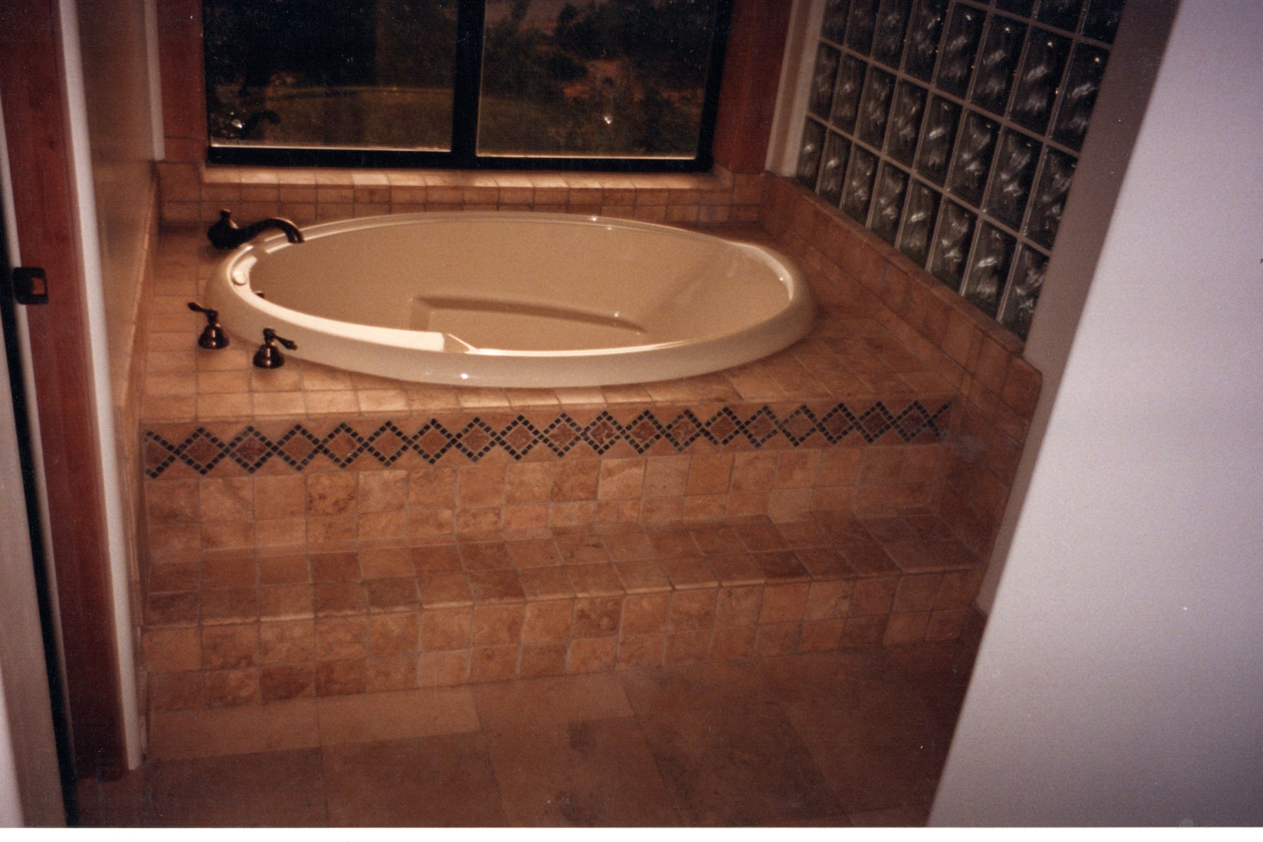 Garden tub set in travertine tile with a mosaic accent band and a glass block wall